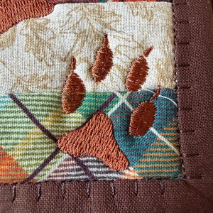 May include: Close-up of a quilted square with embroidered paw prints. The fabric features a plaid pattern in shades of orange, green, and brown, with a cream-coloured section. The paw prints are stitched in a rich brown thread, and the edges are finished with a brown border.
