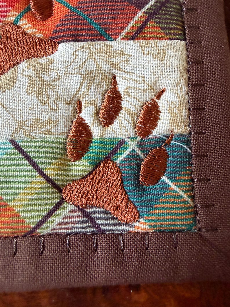 May include: Close-up of a quilted square with embroidered paw prints. The fabric features a plaid pattern in shades of orange, green, and brown, with a cream-coloured section. The paw prints are stitched in a rich brown thread, and the edges are finished with a brown border.
