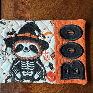 May include: A quilted orange and white Halloween-themed coaster with a black embroidered sloth wearing a witch's hat and skeleton costume. The coaster has the word "BOO" embroidered in black on the orange side.
