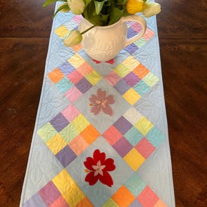 May include: A light blue quilted table runner with a diamond pattern. The runner is decorated with colorful squares and embroidered flowers in pink, red, and purple. A white pitcher with yellow tulips is on top of the runner.
