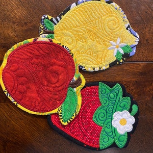 May include: Three quilted fabric pot holders shaped like a lemon, apple, and strawberry. The lemon is yellow with a green stem and leaves. The apple is red with a green stem and leaves. The strawberry is red with green leaves and a white flower.