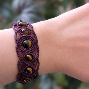 Peut inclure: Bracelet en macramé violet foncé orné de pierres d'œil de tigre polies. Le design tissé du bracelet met en valeur les variations naturelles de couleur des pierres, créant un accessoire unique.