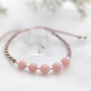 Peut inclure: Un bracelet délicat composé d'un cordon brun clair et de perles roses. Le bracelet est noué et présente un design simple.