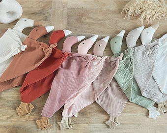 May include: A collection of eight soft goose-shaped security blankets arranged in a row. Each features a plush goose head with a knotted fabric body in various colours including white, rust, red, pink, dusty rose, sage green, and light grey.