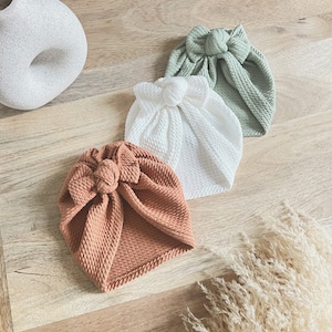 May include: Three turban headbands in different colors: rust, white, and sage green. The headbands are made of a textured fabric and have a knotted bow on top.