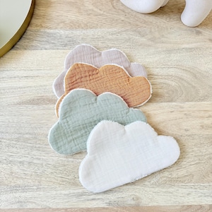 May include: A stack of four cloud-shaped fabric items in pastel colors: light pink, orange, sage green, and white. The items are made of a textured material and are arranged on a wooden surface.