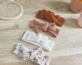 May include: Four headbands in various colors and patterns are displayed on a wooden surface. The headbands include a textured orange, a pink, a white, and a floral print. Each headband features a knotted bow design.
