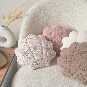 May include: Three decorative pillows in the shape of seashells. The pillows are made of soft, fluffy fabric. One pillow is pink, one is white, and one is brown with a floral pattern.