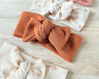 May include: An assortment of baby headbands in various colors and patterns. The headbands include a ribbed rust-colored one, a blush pink one, and two cream-colored ones with floral prints. The headbands are knotted at the top.
