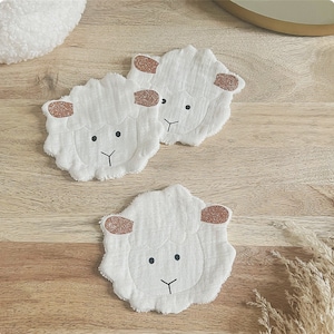 Op de afbeelding: Drie witte stoffen schapen met bruine oren en zwarte ogen. De schapen liggen op een lichtbruin houten oppervlak.