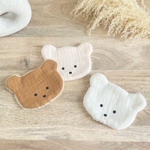 Può includere: Tre lavette per bambini a forma di orso, color crema, bianco e marrone. Ogni lavetta ha naso e occhi neri cuciti. Le lavette sono realizzate in un tessuto morbido e strutturato e sono disposte su una superficie di legno.