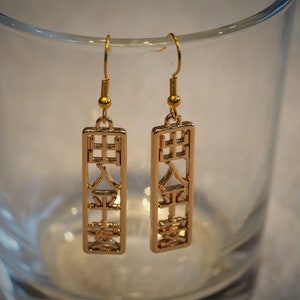Könnte beinhalten: Ein Paar goldfarbene, rechteckige Ohrringe mit japanischen Schriftzeichen. Die Ohrringe hängen an goldfarbenen Haken und werden vor einem klaren Glas präsentiert.