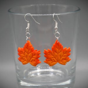 May include: A pair of orange and yellow maple leaf earrings. The earrings are made of a material that resembles clay or plastic. The leaves are detailed with veins and hang from silver-coloured hooks.