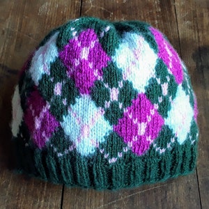 May include: A knitted beanie hat with a green brim and a pattern of pink, white, and light blue argyle diamonds. The hat is made of a soft, textured yarn and is suitable for cold weather.