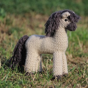 Puede incluir: Un juguete de caballo de punto gris y negro con una cola y una crin largas. El juguete está sentado en un campo de hierba verde.