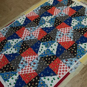 May include: A colourful patchwork quilt with a geometric pattern. The quilt is made of various fabrics featuring cartoon characters and different patterns. The quilt is laid out on a white surface.