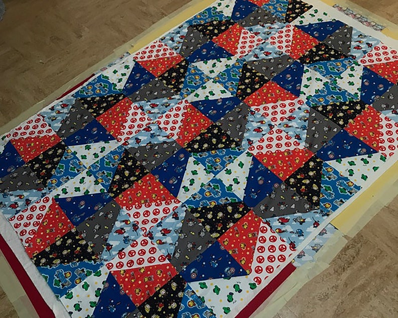 May include: A colourful patchwork quilt with a geometric pattern. The quilt is made of various fabrics featuring cartoon characters and different patterns. The quilt is laid out on a white surface.