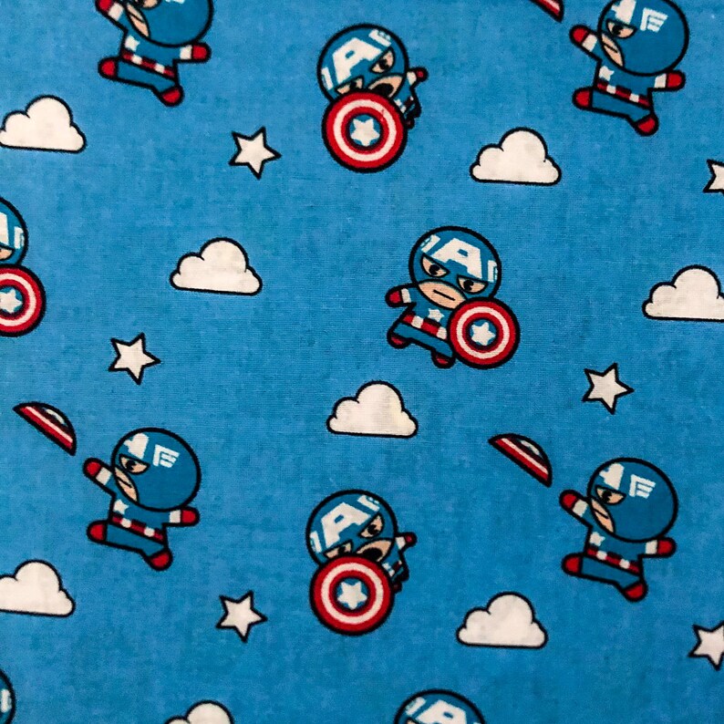 May include: A blue fabric with a repeating pattern of a cartoon superhero wearing a red, white and blue costume. The superhero is holding a red, white and blue shield. The background is a light blue colour with white clouds.