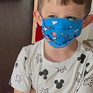 May include: A young child wearing a white t-shirt with a Mickey Mouse pattern and a blue face mask with a colourful design.