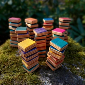 Può includere: Diverse pile di piccole tessere quadrate in legno realizzate in compensato stratificato. Ogni tessera presenta una superficie superiore dai colori vivaci in tonalità giallo, blu, turchese, viola, rosa e arancione, con bordi in legno naturale.