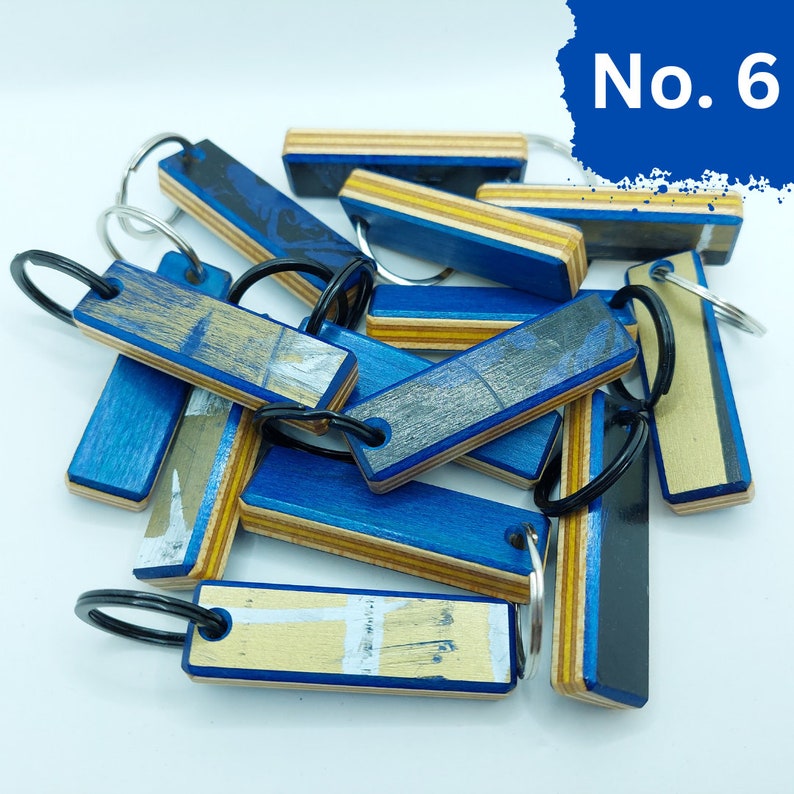 May include: A group of 10 blue and yellow keychains made from recycled skateboard decks. Each keychain has a different design and is attached to a silver key ring. The keychains are arranged in a pile on a white surface. No. 6