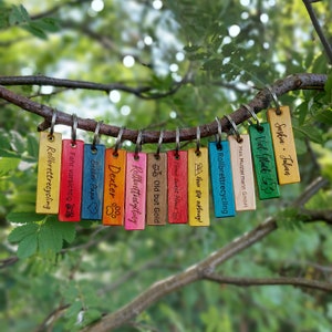 May include: A set of 11 colorful wooden keychains with various phrases and designs. The keychains feature phrases like "Rollbrettrecycling", "Old but Gold", "Never stop exploring", and "Home sweet Home".