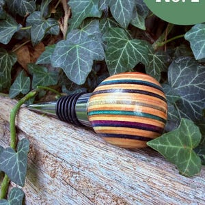 May include: A bottle stopper featuring a spherical handle crafted from multi-colored laminated wood layers in shades of natural tan, teal, purple, and black, attached to a tapered metal base with black rubber rings for a secure seal.