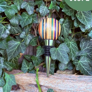 May include: A bottle stopper featuring a spherical top made of multi-colored laminated wood strips in shades of red, orange, yellow, green, blue, and black. The stopper has a silver-toned metal base with black rubber rings for a secure seal.