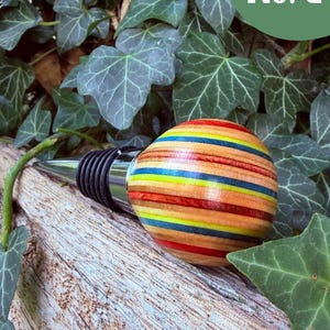 May include: A bottle stopper featuring a spherical handle crafted from layers of multi-colored wood in shades of red, yellow, blue, and natural tan. The stopper has a polished chrome metal base with black rubber rings for a secure fit.