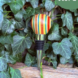 May include: A bottle stopper featuring a rounded top made of multi-colored, vertically striped wood in shades of red, yellow, and blue. The stopper has a polished silver-tone metal base with black rubber rings for a secure fit. Text reads No. 2.