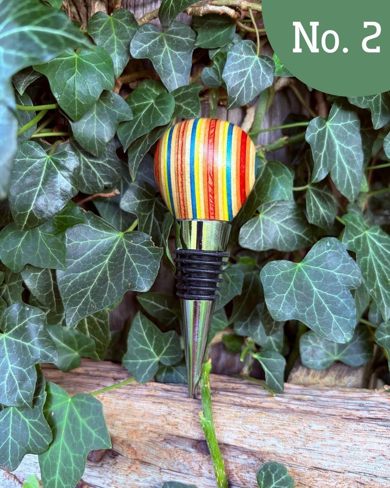 May include: A bottle stopper featuring a rounded top made of multi-colored, vertically striped wood in shades of red, yellow, and blue. The stopper has a polished silver-tone metal base with black rubber rings for a secure fit. Text reads No. 2.