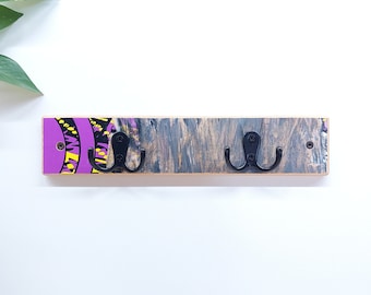 May include: A wooden wall-mounted coat rack with two double hooks. The rack is painted with a purple, yellow, and black abstract design.