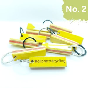 May include: A close-up of a yellow wooden keychain with the text "Rollbrettrecycling" engraved on it. The keychain is made from recycled skateboard wood and has a white stripe down the center. The keychain is attached to a silver key ring.