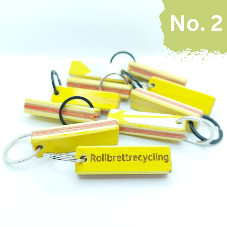 May include: A close-up of a yellow wooden keychain with the text "Rollbrettrecycling" engraved on it. The keychain is made from recycled skateboard wood and has a white stripe down the center. The keychain is attached to a silver key ring.