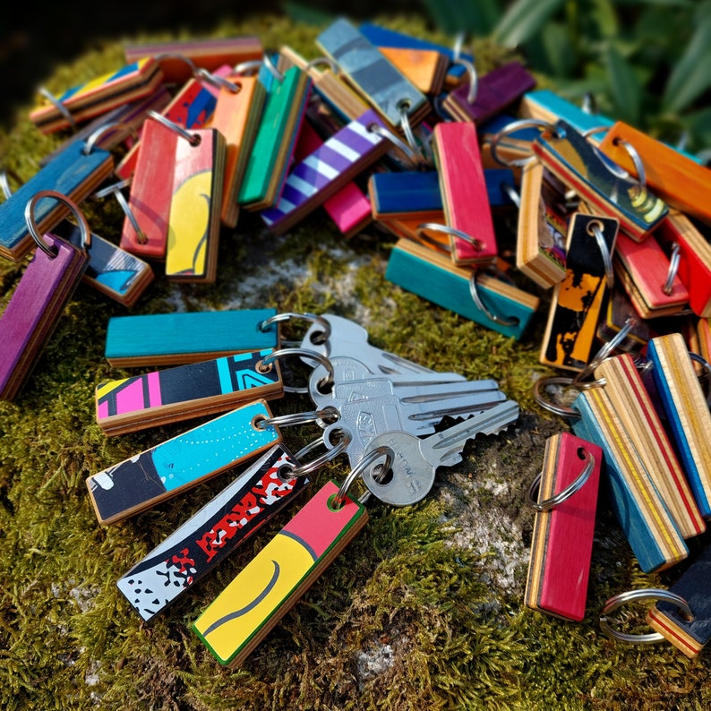 May include: A collection of colorful keychains made from recycled skateboard decks. Each keychain features a unique design and is attached to a silver metal ring. The keychains are arranged on a bed of green moss.