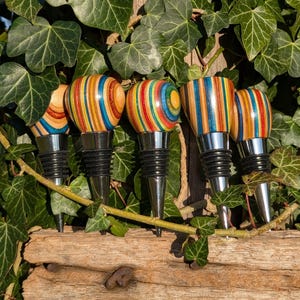 May include: Five bottle stoppers with polished chrome bases and black rubber rings. Each stopper features a unique handle crafted from multi-colored laminated wood, displaying vibrant stripes of blue, orange, red, yellow, and natural wood tones.