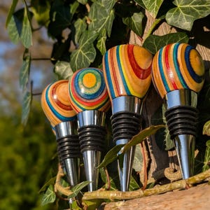 May include: Four bottle stoppers featuring rounded, multi-colored wooden tops with concentric rings of yellow, blue, red, and natural wood grain. Each stopper has a polished silver-tone metal base with black rubber rings for a secure seal.