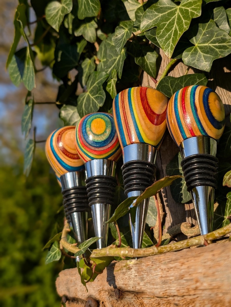 May include: Four bottle stoppers featuring rounded, multi-colored wooden tops with concentric rings of yellow, blue, red, and natural wood grain. Each stopper has a polished silver-tone metal base with black rubber rings for a secure seal.