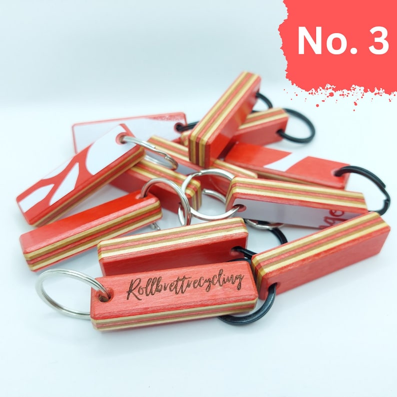 May include: A close-up of a pile of red and white wooden keychains with black metal rings. The keychains are made from recycled skateboards and have the words "Rollbrettrecycling" engraved on one of them.