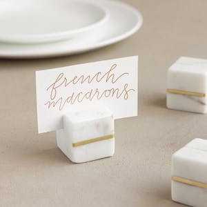 Op de afbeelding: Witte marmeren plaatskaartjeshouders met een gouden accent. De kaarthouder houdt een kaart vast met de tekst "french macarons".