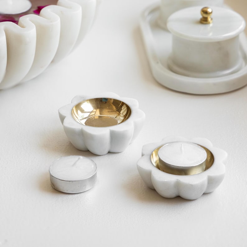 以下が含まれることがあります： 金色のアクセントが付いた白い大理石のキャンドルホルダーが2つ。それぞれのホルダーは波打つ縁取りと中央に小さな丸い窪みがあります。1つのホルダーには白いティーライトキャンドルが入っています。