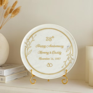 May include: A white decorative plate with gold lettering and accents, commemorating a 38th anniversary. The plate reads "Happy Anniversary Mommy & Daddy November 14, 1987" and is displayed on a gold stand.