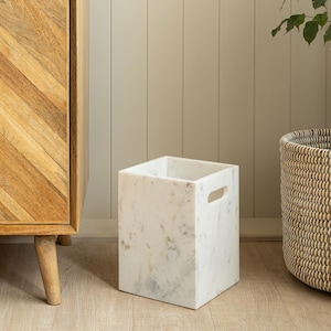 May include: A square, white marble waste bin with subtle grey veining. The bin has two rectangular cut-out handles. It sits on a light wood floor. The background includes a wooden cabinet and a woven basket.