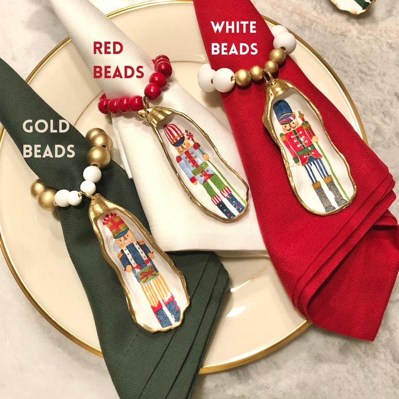 May include: Three Christmas napkin rings with a nutcracker design. Each ring is made of a painted oyster shell with gold trim and a different colored beaded garland: red, white, and gold.