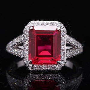 Può includere: Un anello d'argento con una grande gemma rossa vibrante di forma rettangolare, circondata da un alone di gemme chiare più piccole. Il gambo dell'anello si divide in due, ciascuna con altre gemme chiare, creando un effetto scintillante.
