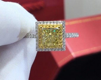May include: A square-cut yellow diamond ring with a halo of smaller diamonds. The ring has a silver band with additional small diamonds. The ring is set against a red background.