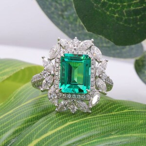 Può includere: Un anello d'argento con una grande gemma verde rettangolare taglio smeraldo, circondata da un alone di pietre più piccole, chiare, a forma di navetta. La fascia è impreziosita da piccole pietre rotonde e chiare. L'anello è su una foglia verde.