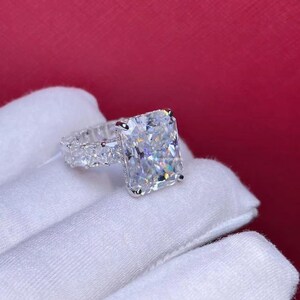 Pode incluir: Um anel de prata com uma grande pedra central de diamante quadrada e transparente. A banda &eacute; adornada com diamantes menores e transparentes.
