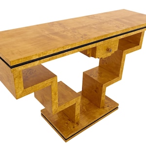 Op de afbeelding: Een lichtgekleurde houten console met een geometrisch ontwerp. De tafel heeft een rechthoekig blad met een zwarte rand en een trapsgewijze basis. De houtnerf is zichtbaar. De tafel is ongeveer 152 cm lang.