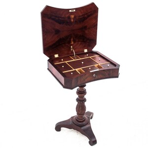 Puede incluir: Una mesa de madera oscura con una tapa con bisagras que se abre para revelar un compartimento con varios cajones pequeños. La mesa tiene una base de pedestal única con un diseño tallado.